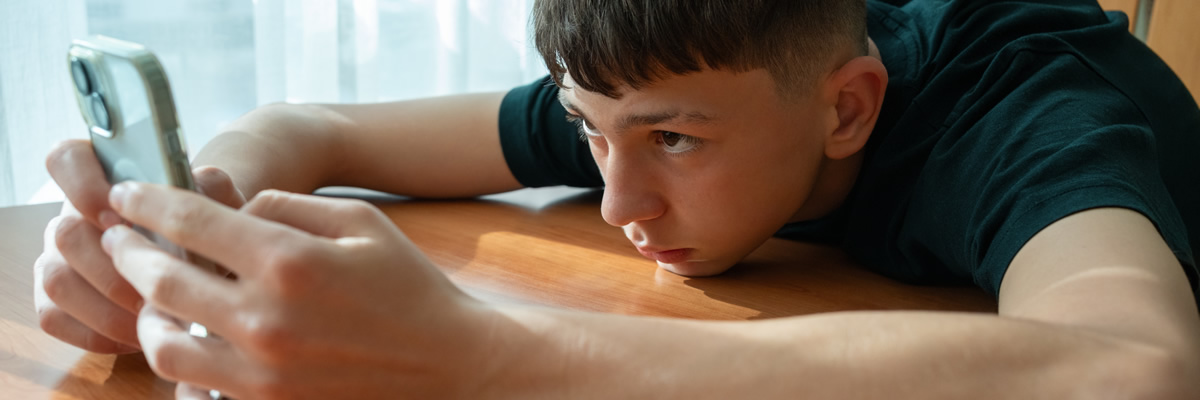 Ein Junge liegt erschöpft auf dem Tisch und starrt intensiv auf sein Smartphone.